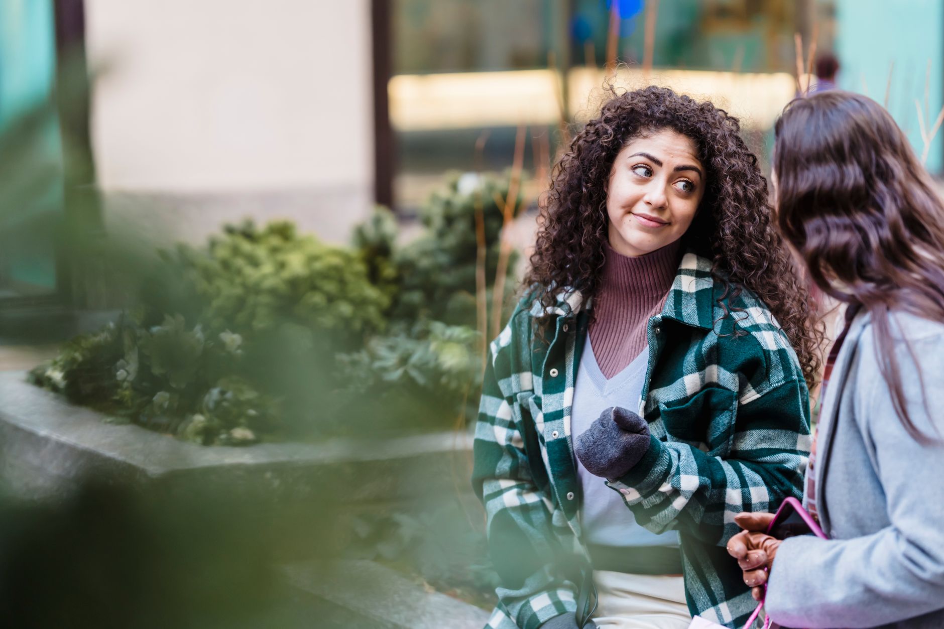Calm ethnic woman with dark curly hair in trendy clothes standing and communicating with friend on street against blurred building in daytime