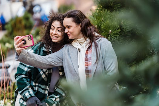 Two women smiling and taking a selfie outdoors near lush green plants in a city garden.