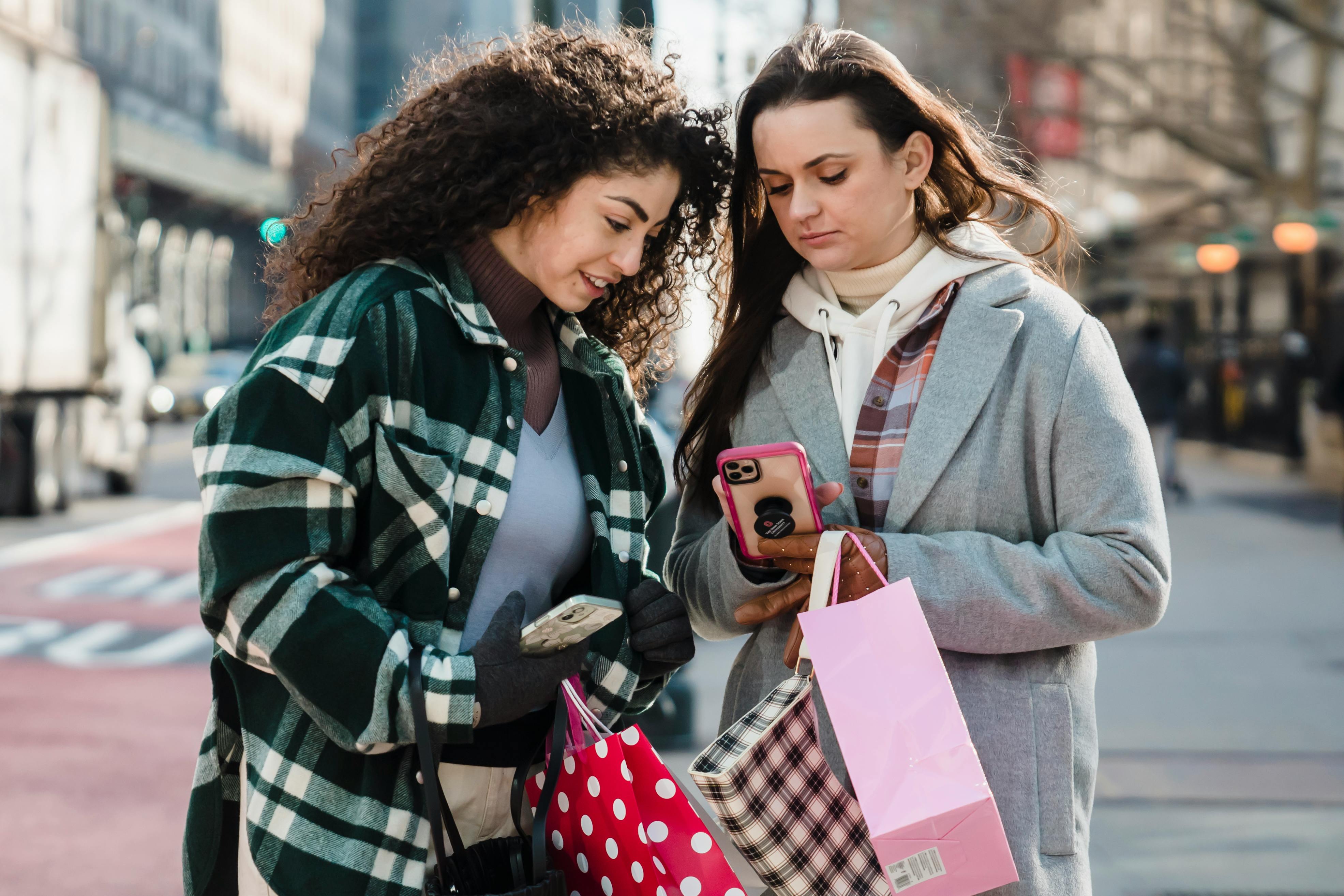 friends standing with shopping bags on street and looking at smartphone