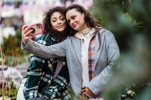Positive young multiracial female best friends in stylish clothes cuddling and smiling while taking selfie on smartphone in city park