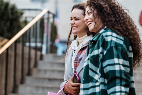 Side view of joyful young multiethnic female best friends with dark hair in stylish outfits smiling happily looking away while standing on city street near staircase in daytime