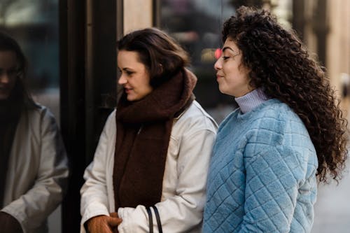 Side view of trendy young multiracial females in stylish warm clothes standing near glass showcase of fashion boutique before shopping