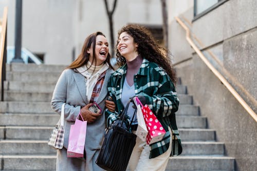 Free Joyful young stylish multiracial female best friends in trendy warm clothes laughing while walking downstairs with shopping bands in hands on city street Stock Photo