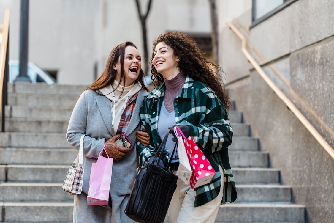 Joyful young stylish multiracial female best friends in trendy warm clothes laughing while walking downstairs with shopping bands in hands on city street
