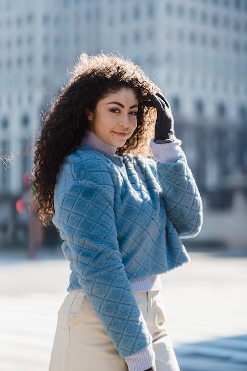 Cheerful young female in gloves and warm fashionable outfit smiling and looking at camera on blurred background of street