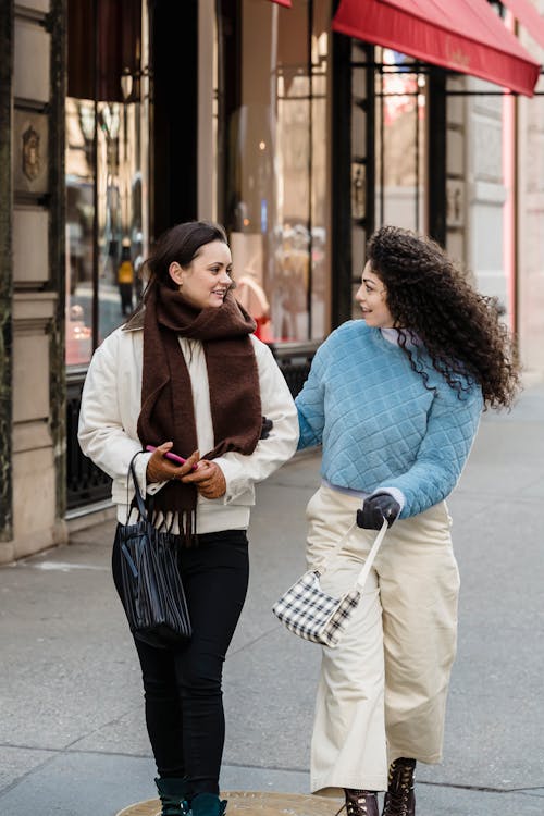 Happy trendy friends in scarf and soft pullover speaking while promenading on contemporary street of city