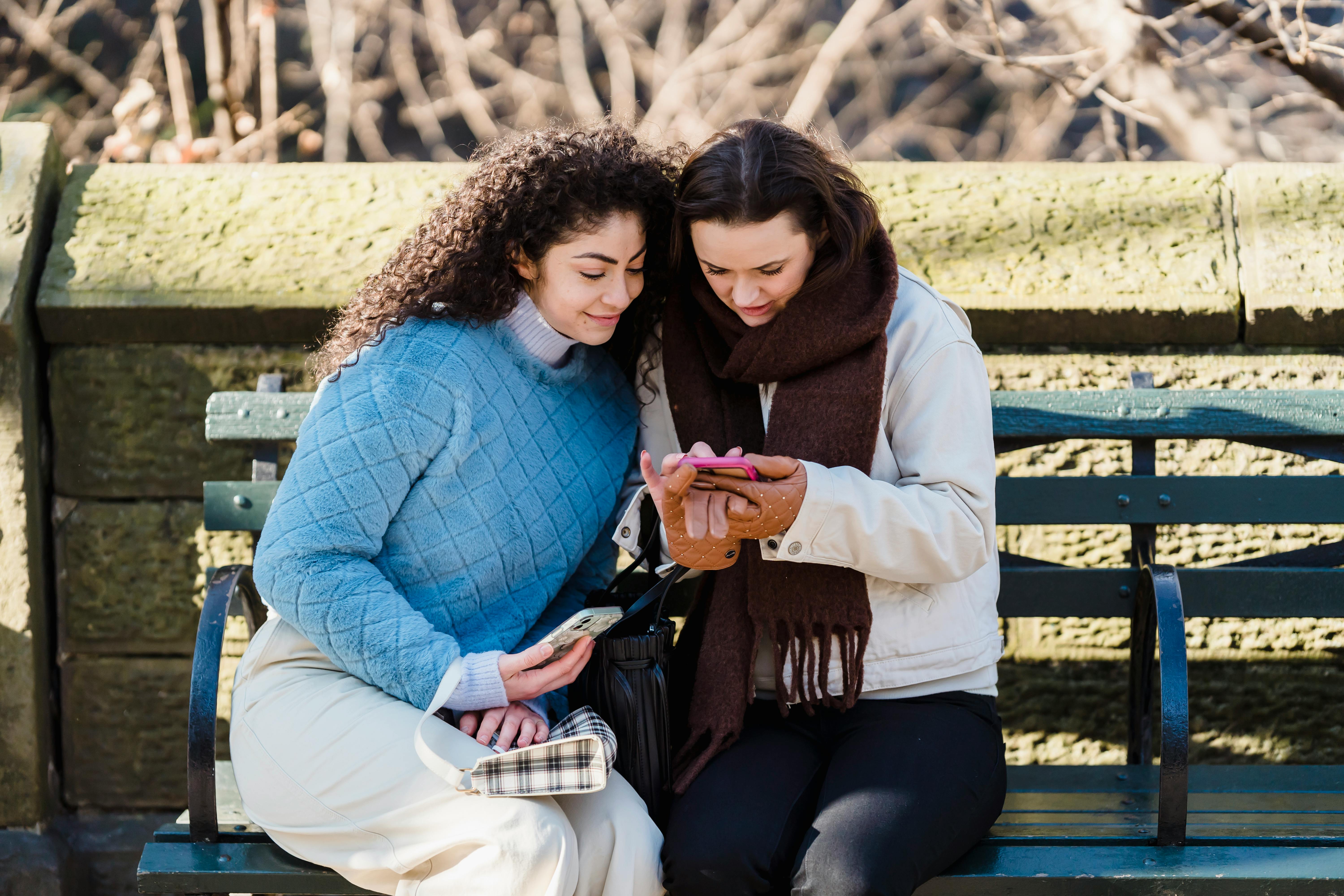 trendy women using smartphone together on bench in park