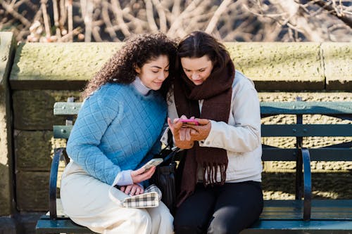 Young happy friends smiling while surfing internet on mobile phone in sunny park in daytime