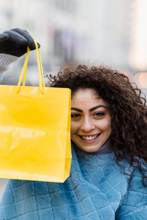 Free Cheerful young woman with curly hair holding a bright yellow shopping bag outdoors, showcasing joy and shopping excitement. Stock Photo