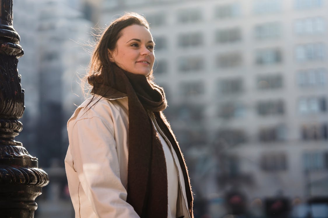Glad female in trendy brown scarf smiling and looking away on blurred background of urban building