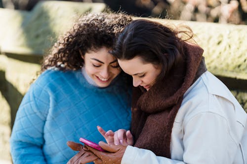 Free Cheerful women smiling and looking at screen while surfing internet on mobile phone in park in daytime Stock Photo