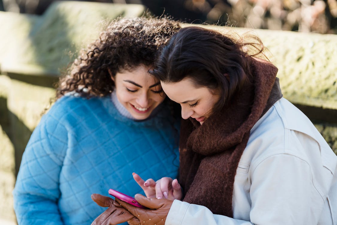 Cheerful women smiling and looking at screen while surfing internet on mobile phone in park in daytime