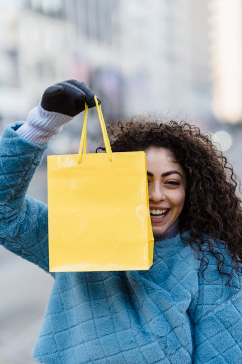 Free Cheerful young female squinting and smiling while demonstrating yellow gift bag hanging in hand Stock Photo