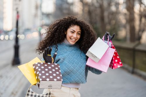 Free Happy customer with many different colorful gift bags in trendy sweater smiling on blurred background of street Stock Photo