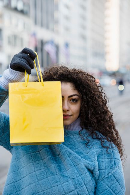 Young female with long curly hair showing yellow gift bag in front of face on blurred background of urban street