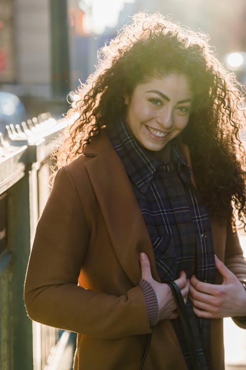 Happy young female with long hair in trendy clothes smiling and looking at camera in back lit