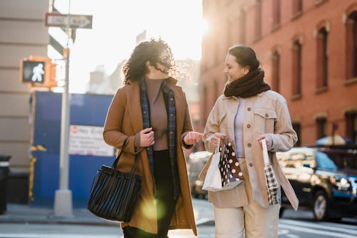 Positive young stylish ladies with long dark hairs in trendy warm outfits crossing road while spending time together in city on sunny day