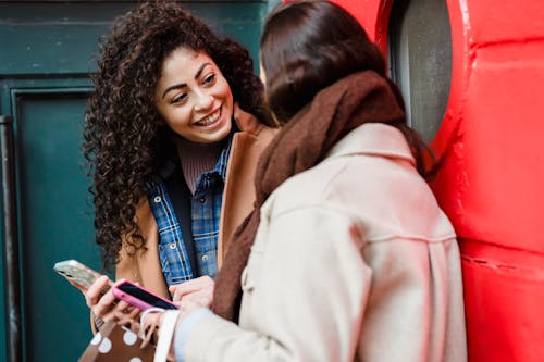 Smiling young multiracial ladies browsing smartphones and communicating on street