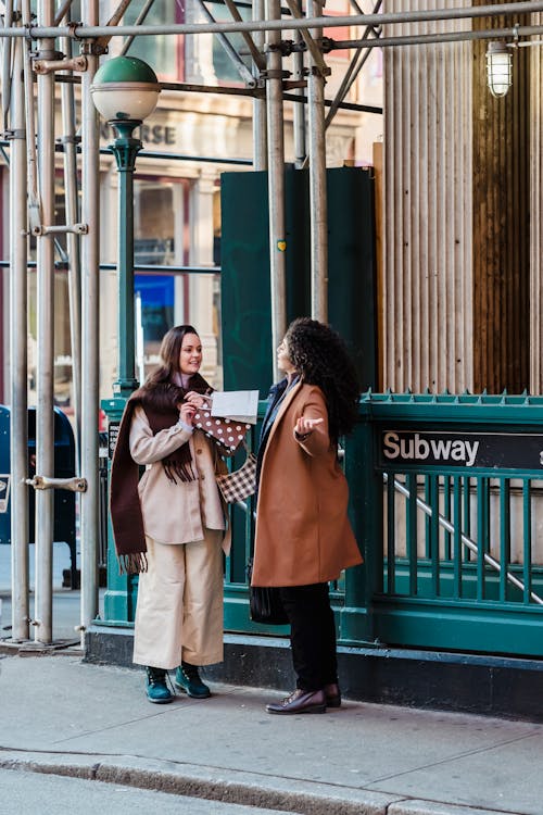 Full body of positive young female colleagues in stylish clothes standing on street near entrance of subway and communicating during break