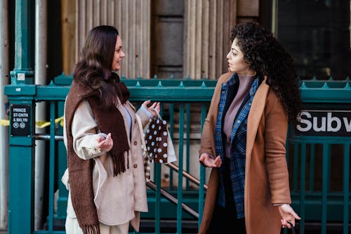 Side view of young stylish multiracial female friends in fashionable outfits standing on street and gossiping near subway station