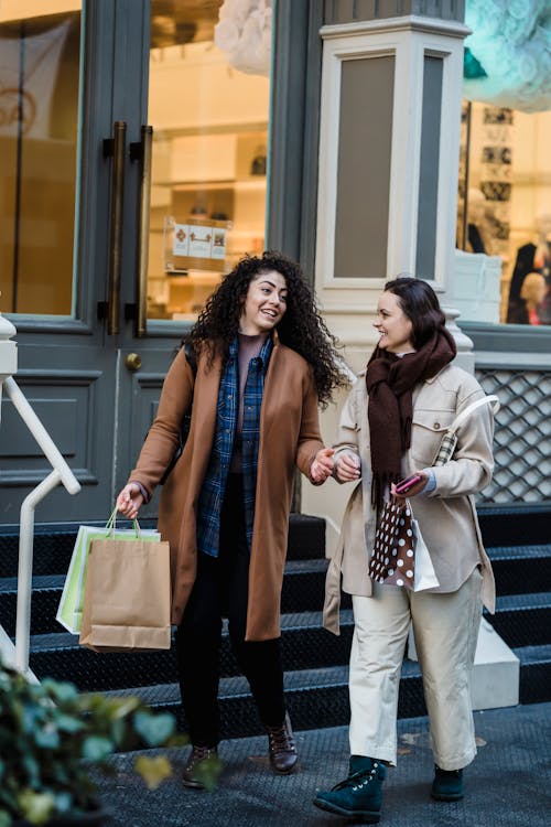 Full body of delighted young multiethnic ladies with dark hair in stylish outfits smiling and talking while walking on street near entrance of fashion boutique after shopping