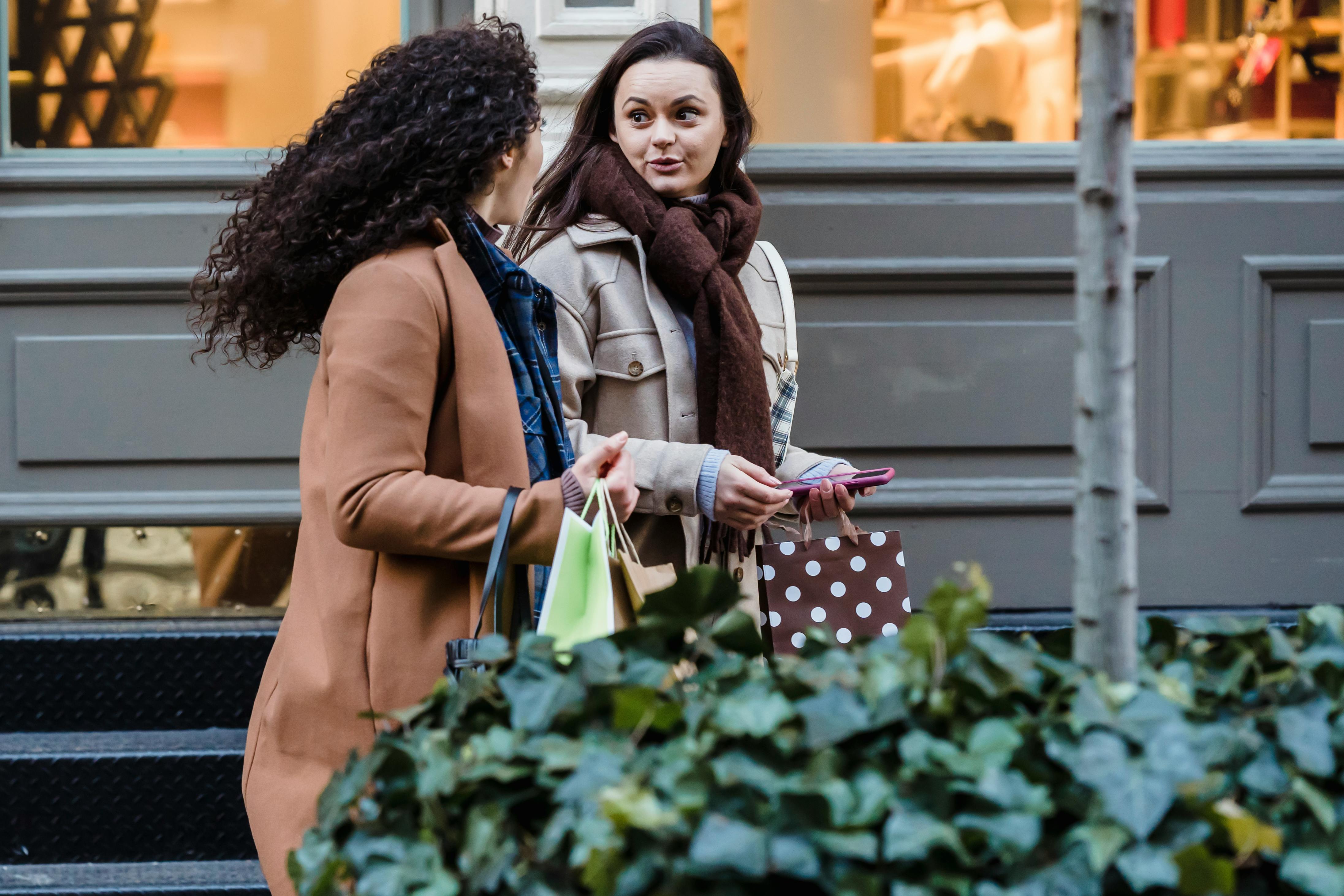 amazed young women with shopping bags chatting while strolling on sidewalk