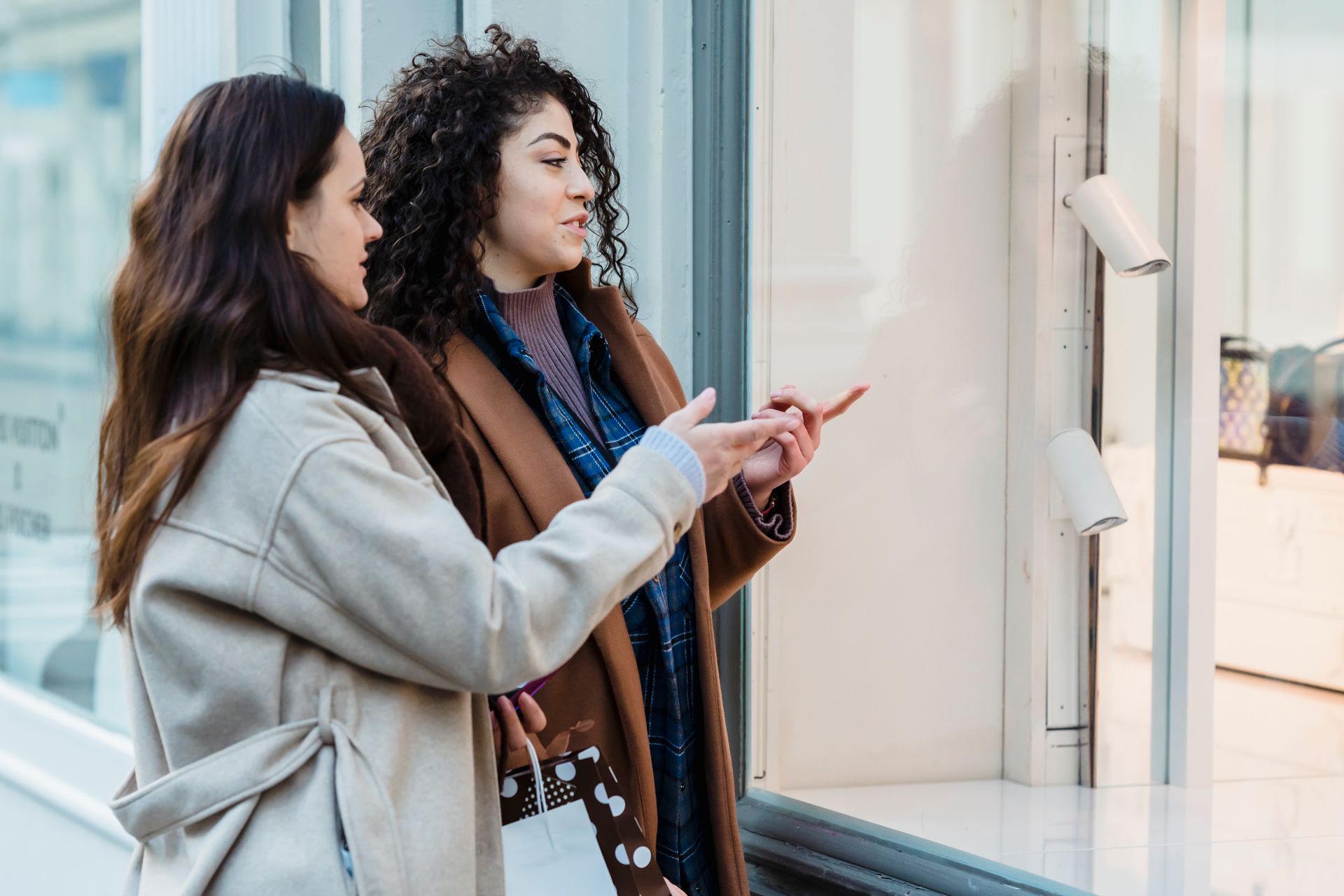 Side view of positive young multiracial female friends standing on city street and pointing on showcase of fashion boutique during shopping