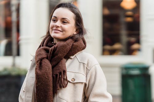 Confident young female with long brown hair in stylish coat and scarf smiling while standing on city street in daytime