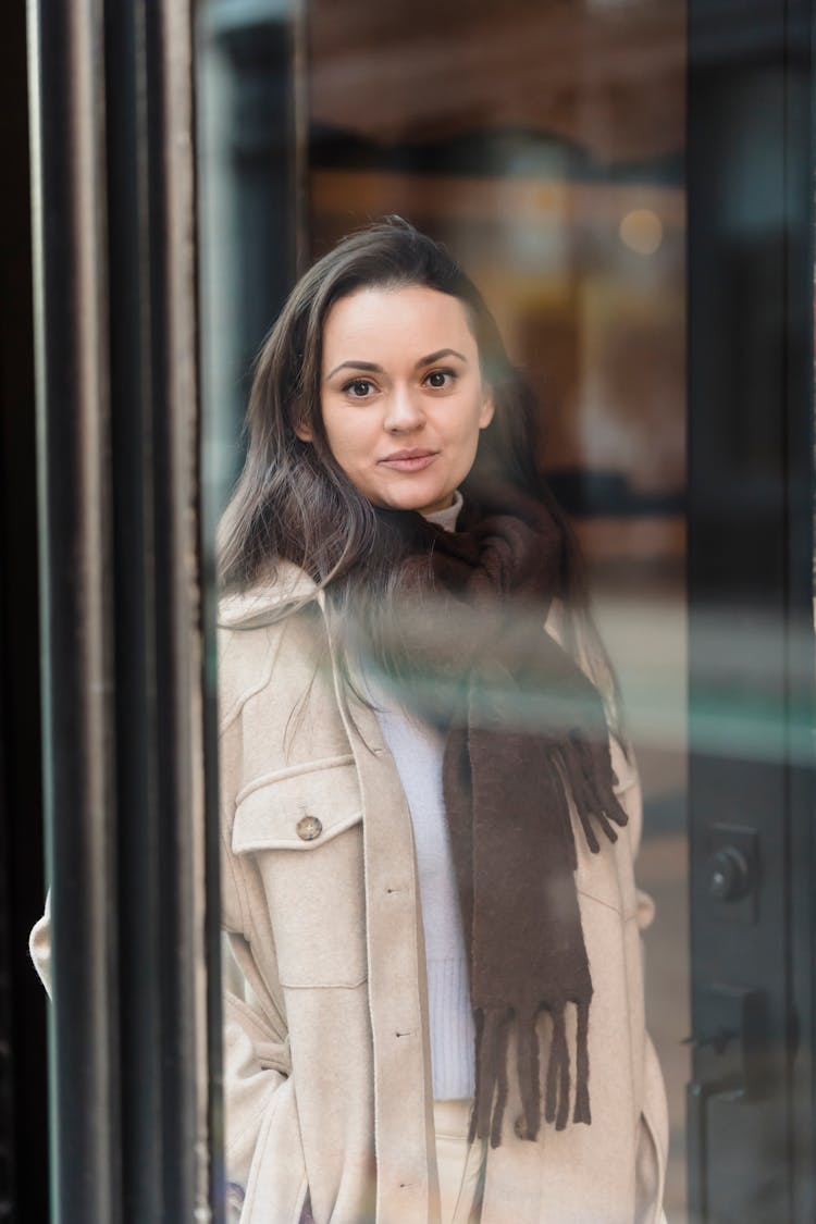 Smiling Young Woman Standing On Street And Looking At Camera Through Glass Door