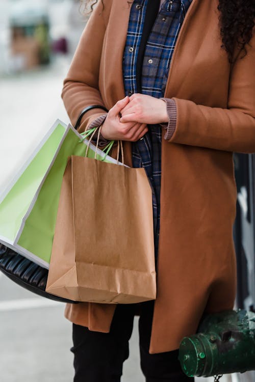 Free Crop unrecognizable female buyer in elegant coat carrying paper bags while standing on city street after shopping Stock Photo