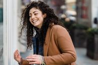 Cheerful young ethnic female with long curly hair in stylish clothes laughing while standing on city street during weekend