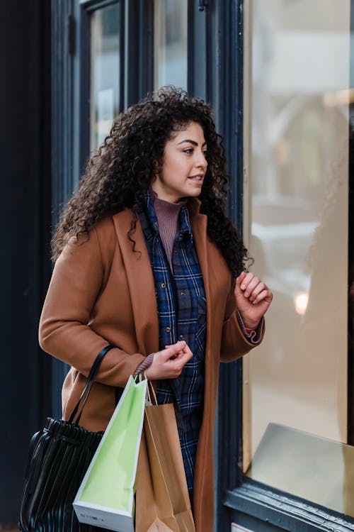 Confident young ethnic woman with dark curly hair in casual outfit looking at store showcase while walking on street with shopping bags