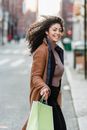 Side view of cheerful ethnic young female shopper with flying curly hair carrying paper shopping bags running on city road