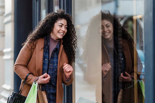 Laughing ethnic female in coat and casual clothes standing with paper bags near showcase after shopping