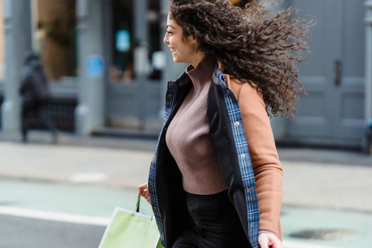 Positive young ethnic female with flowing hair carrying shopping paper bags while quickly crossing road and looking away