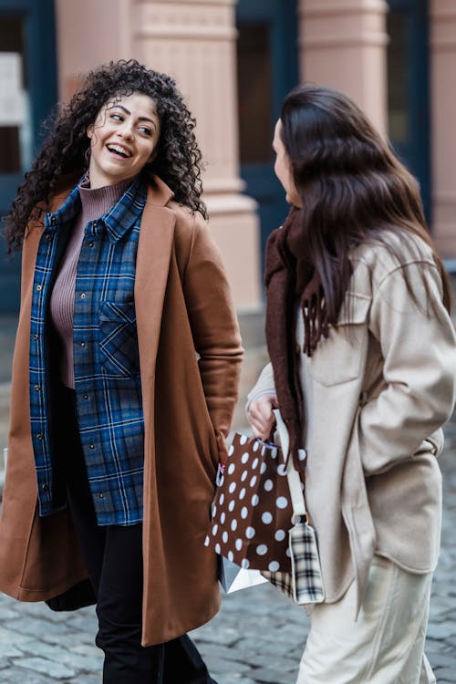 Glad ethnic female wearing coat walking near friend carrying gift bag on paved city street