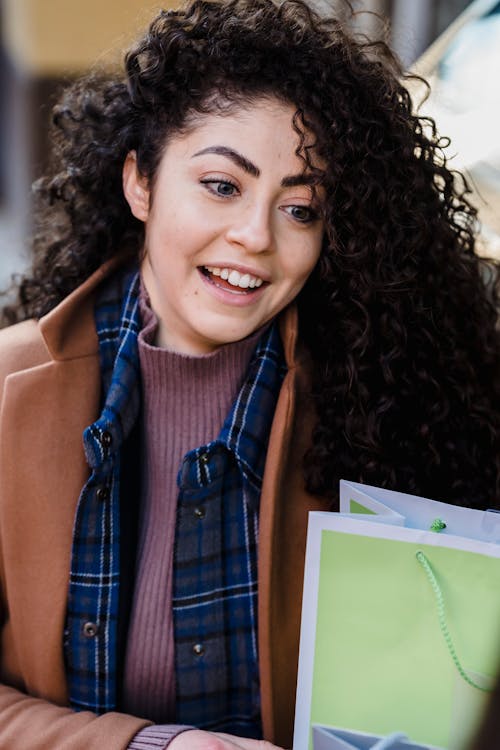 Free High angle of smiling ethnic female with curly hair wearing outerwear sitting with paper bag and looking away Stock Photo