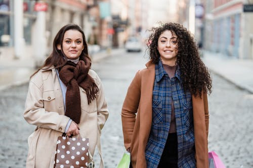 Smiling multiracial girlfriends with hands in pockets standing on paved street with gift bags and looking at camera