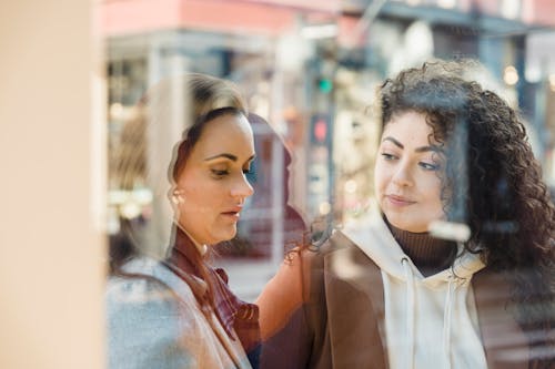 Through glass of focused multiracial female friends standing near window of store while choosing goods on sunny street in city