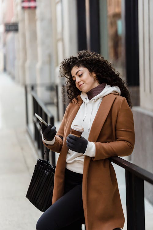 Free Side view of ethnic female browsing cellphone while standing near metal railing with takeaway coffee on street with building on blurred background Stock Photo