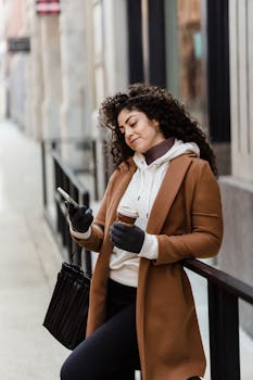 Side view of ethnic female browsing cellphone while standing near metal railing with takeaway coffee on street with building on blurred background