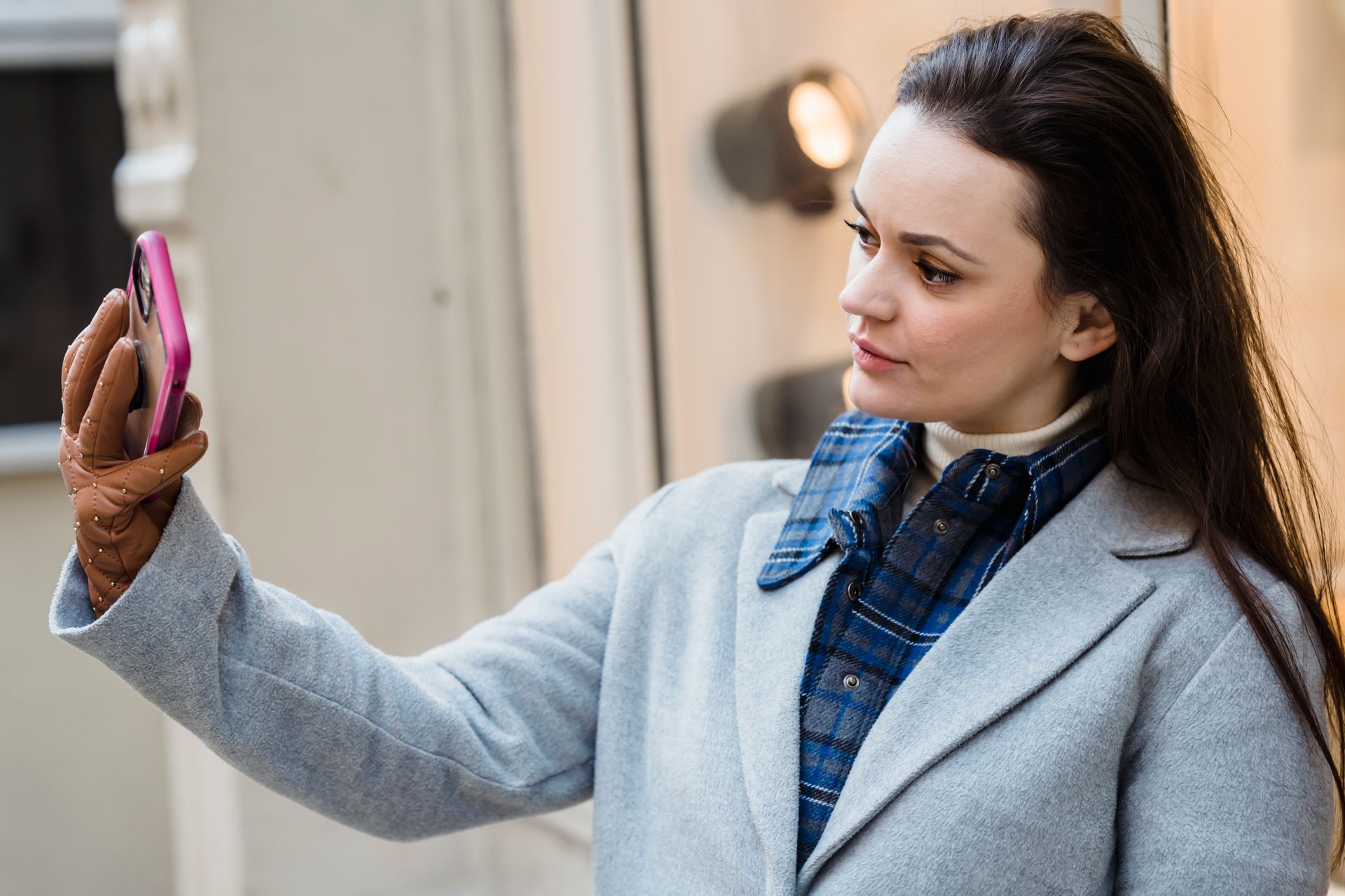Side view of serious female in warm clothes taking self portrait on smartphone while standing on street near building in city