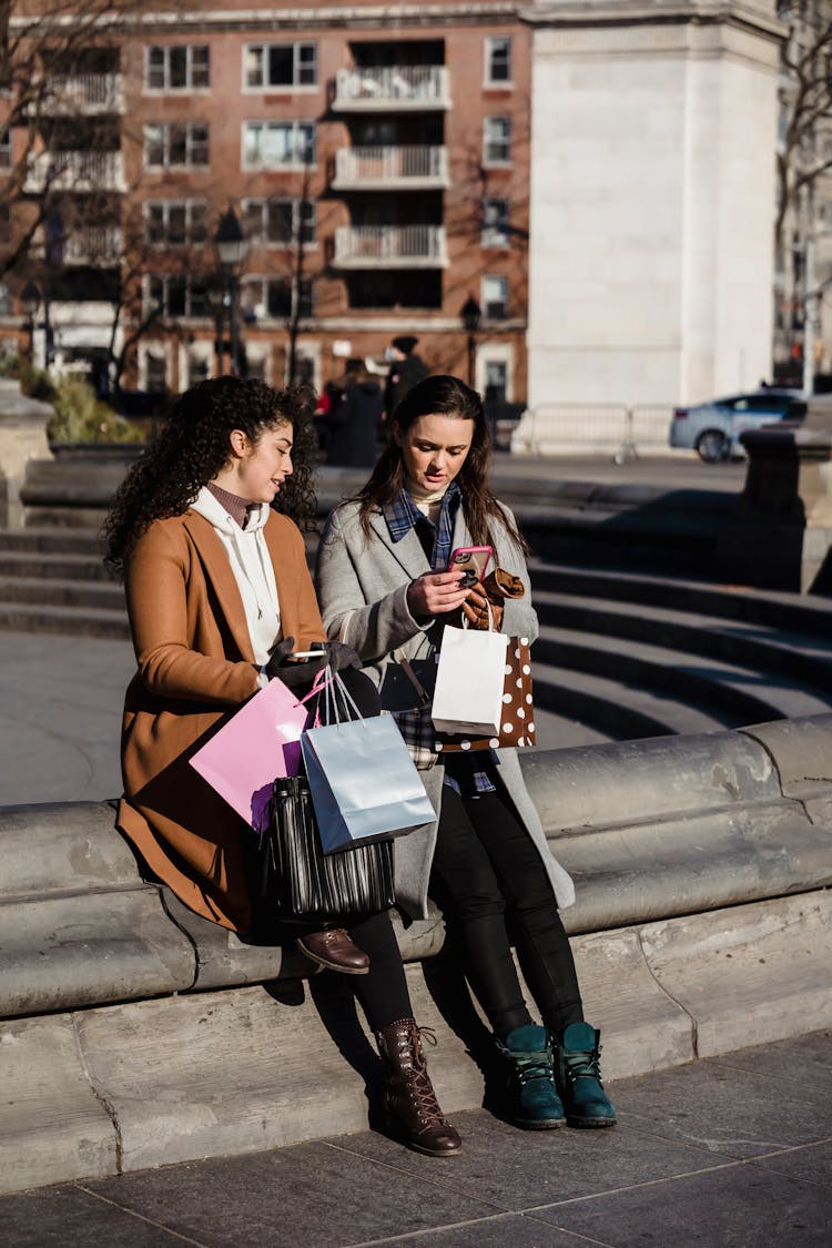 Content Women With Shopping Bags And Smartphone Sitting On Street