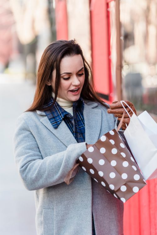 Free Excited young female wearing blue coat looking in shopping bags while standing on street on sunny cold weather Stock Photo
