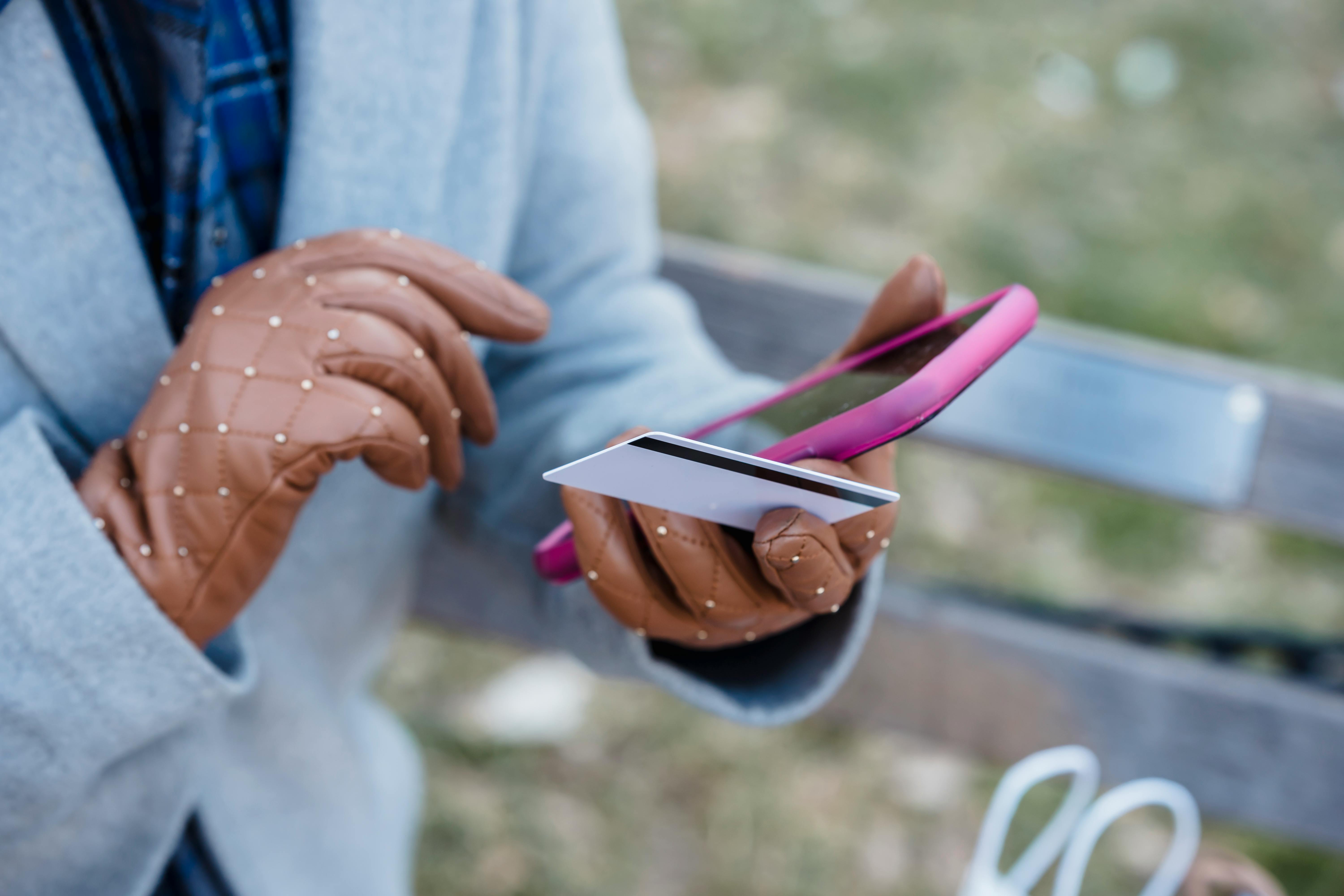 Free Crop anonymous female in blue coat and leather gloves with credit card making online purchase via smartphone while sitting on bench in city park Stock Photo