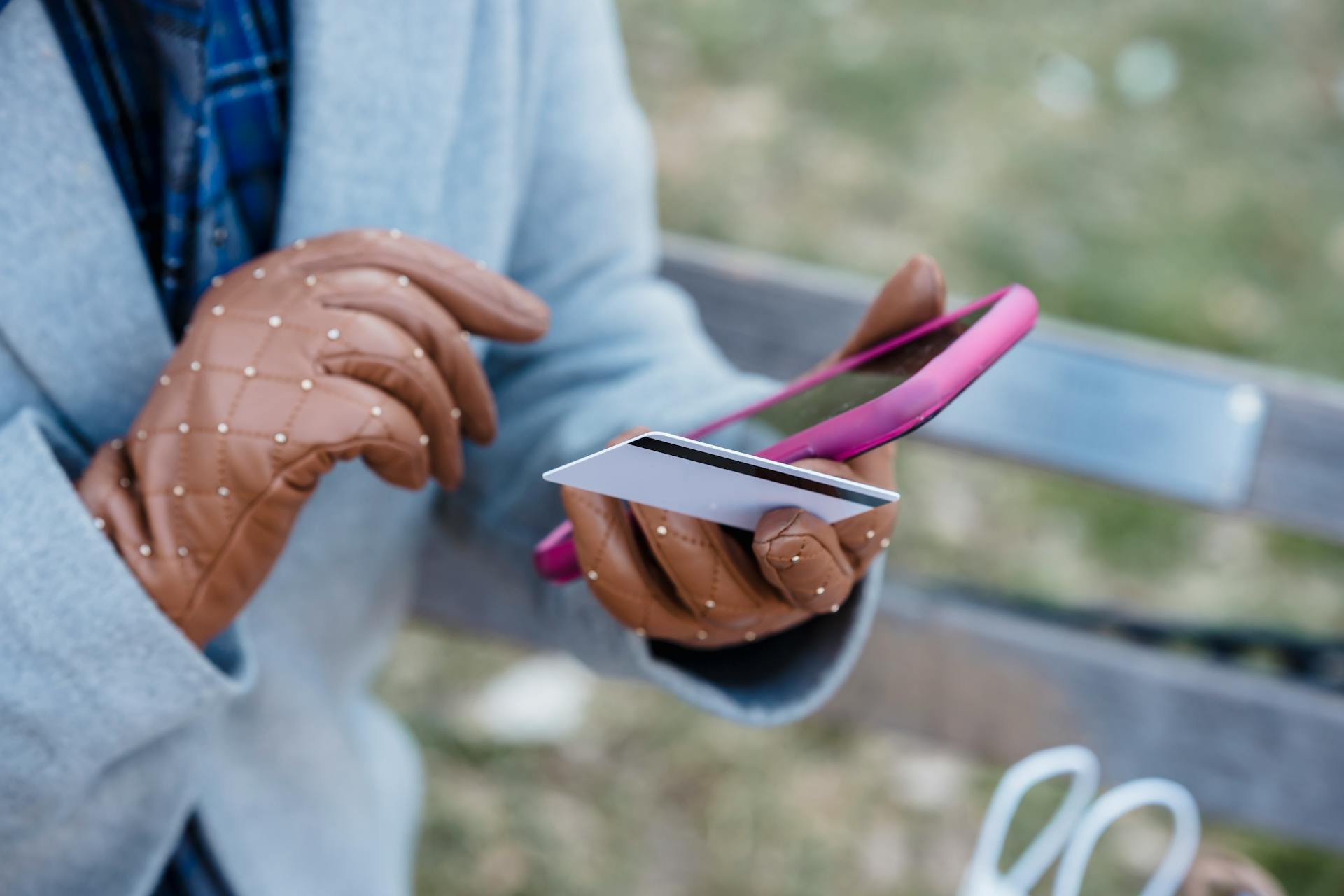 Crop anonymous female in blue coat and leather gloves with credit card making online purchase via smartphone while sitting on bench in city park