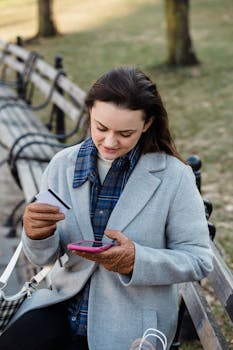 Content young female in blue coat making online payment with smartphone while sitting on bench in park on early spring day
