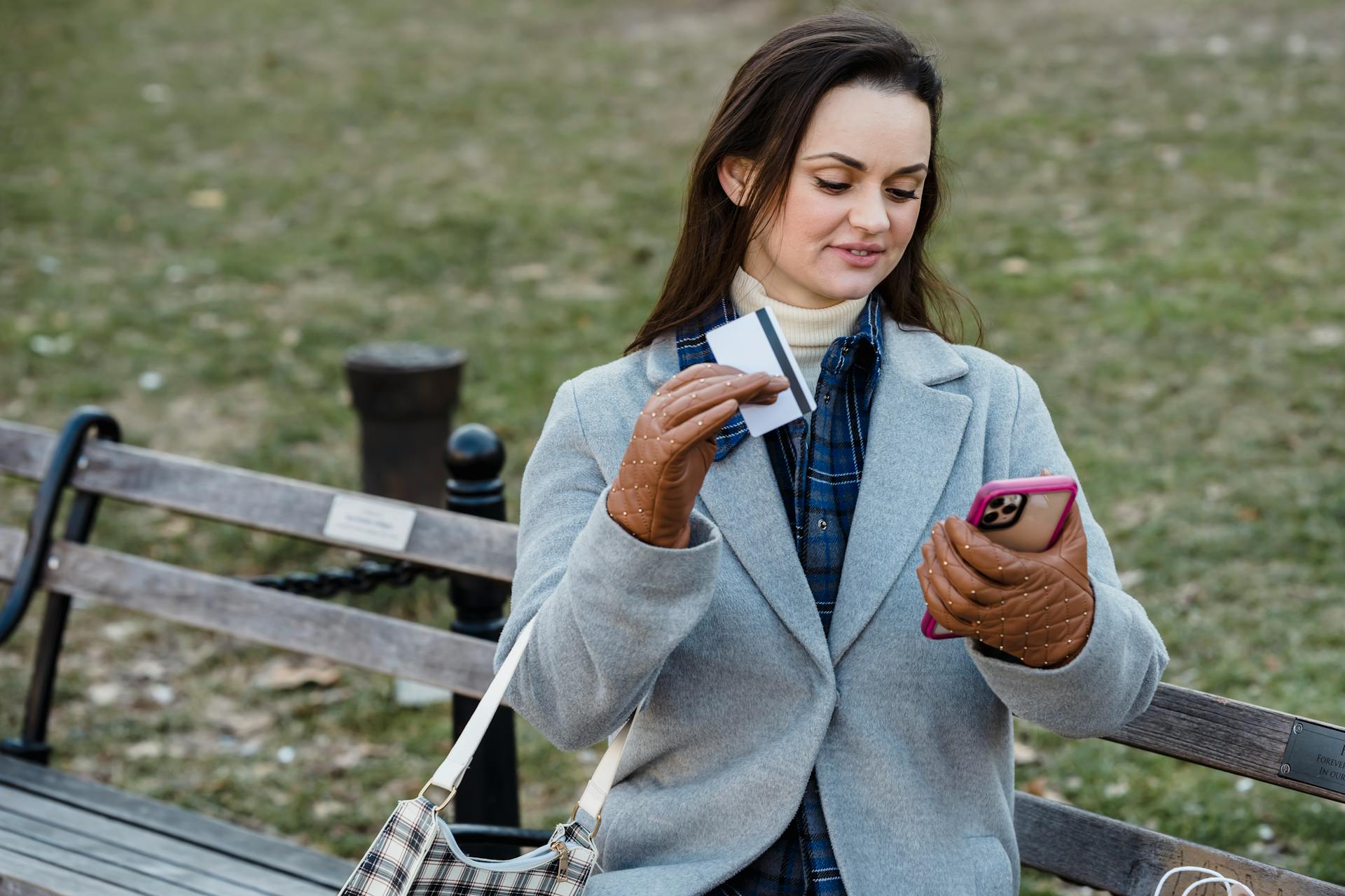 Young woman sitting on bench using smartphone and credit card for online shopping outdoors.