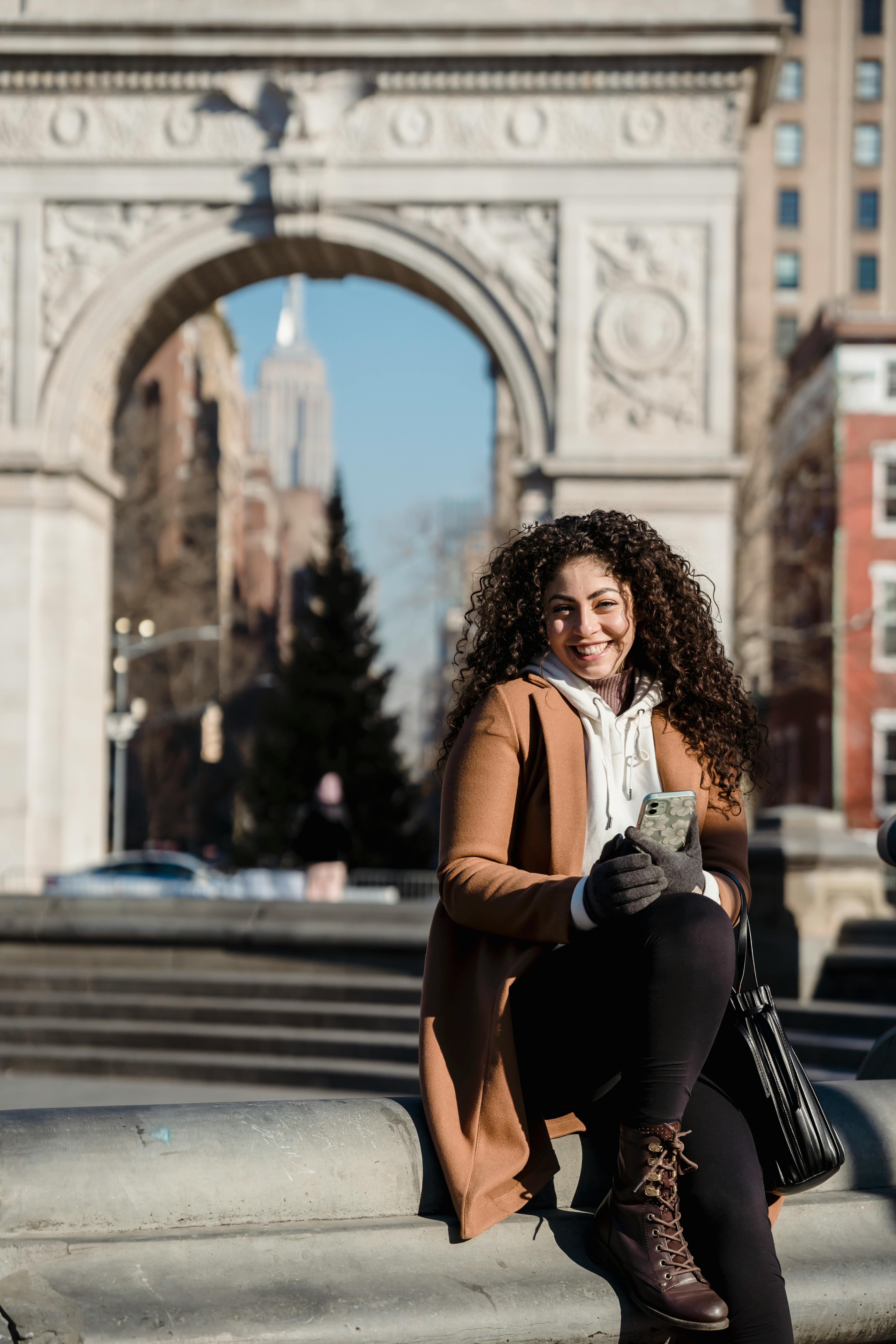 happy woman browsing smartphone and sitting on stone border on street