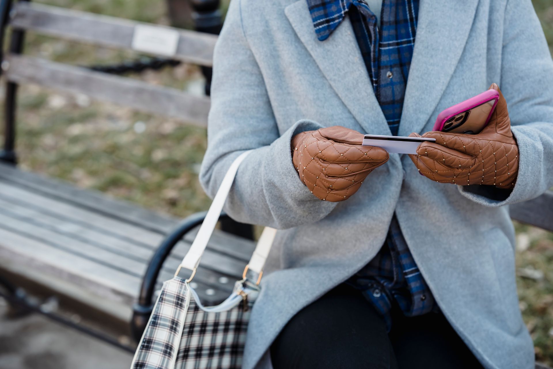 A woman using her smartphone for a transaction while sitting on a park bench wearing gloves and a coat.